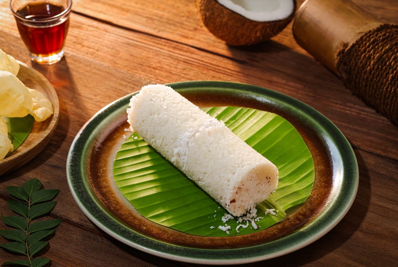 A white rice puttu with grated coconut on a banana leaf, with a coconut and a glass of tea beside it